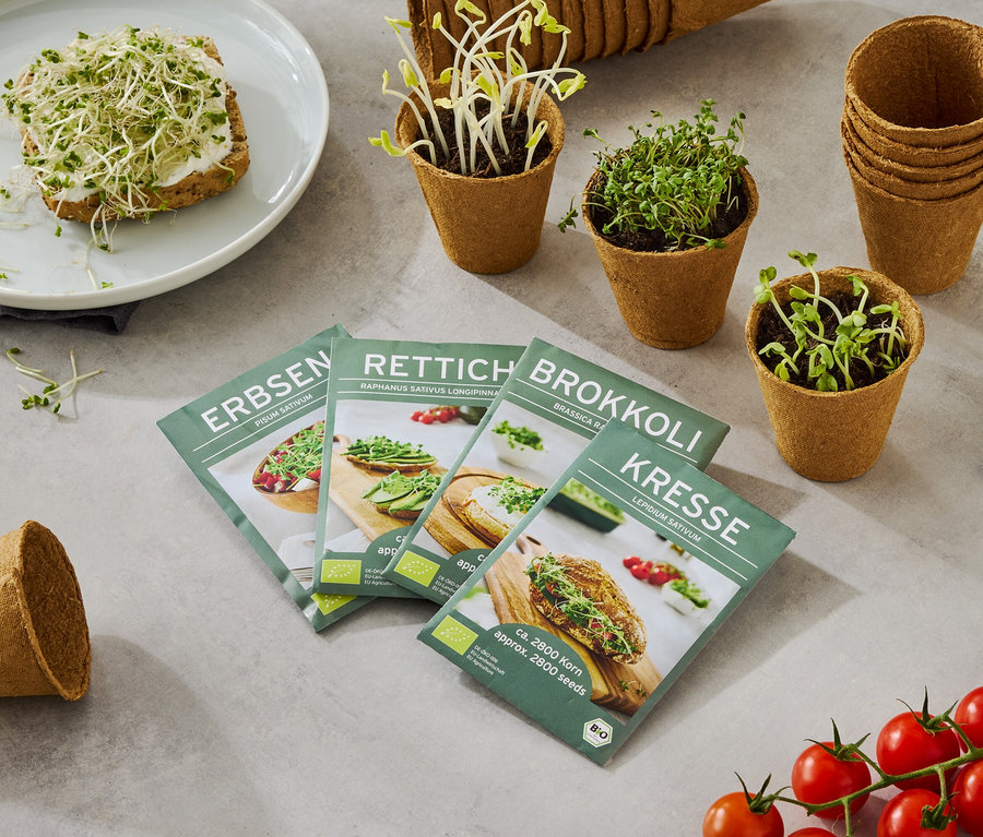 Ein Teller mit einem mit Sprossen belegten Brot, zusammen mit vier Samentüten (Erbsen, Rettich, Brokkoli, Kresse), kleinen Töpfen mit wachsenden Sprossen und einer Rispe Tomaten.