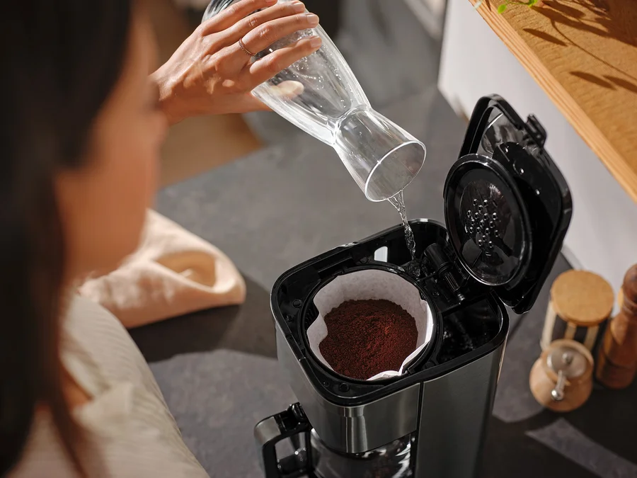 Frau gießt Wasser aus einer Glaskaraffe in eine Kaffeemaschine.
