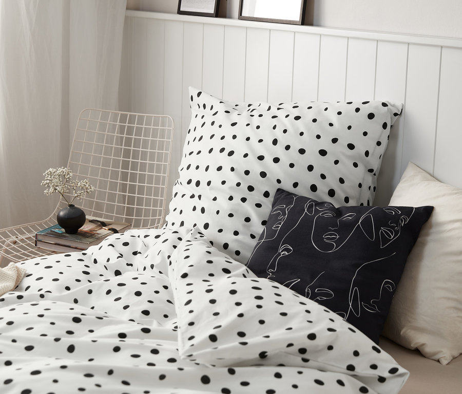 Bettwäsche mit schwarzen Punkten auf einem Bett mit einem schwarz-weißen Kissen und einer schwarzen Vase mit Blumen auf einem weißen Stuhl.