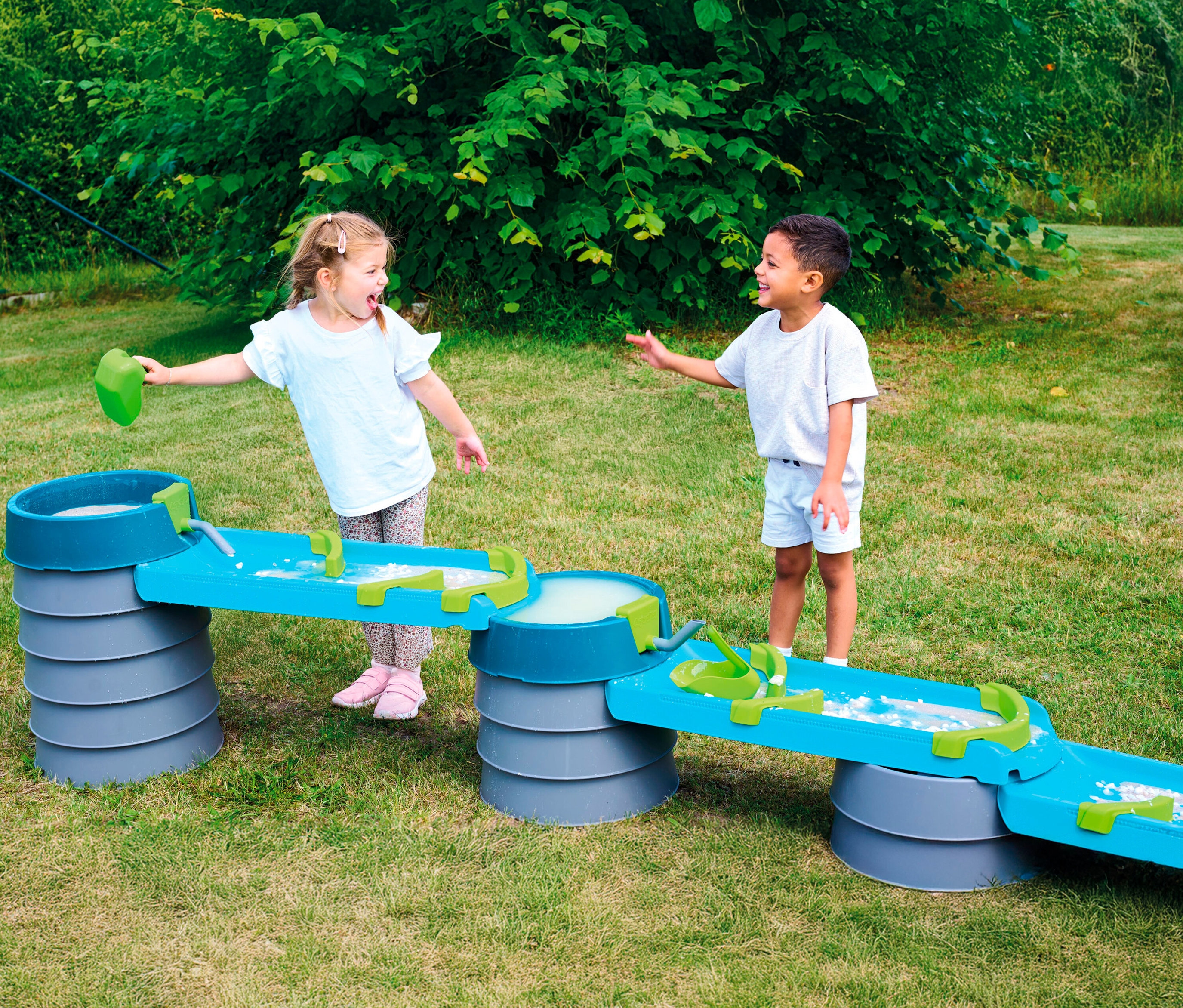 Zwei Kinder spielen mit der AquaPlay »Mudway STREAM« Matschbahn im Garten. Ein Mädchen gießt Wasser in die Bahn, ein Junge steht daneben.