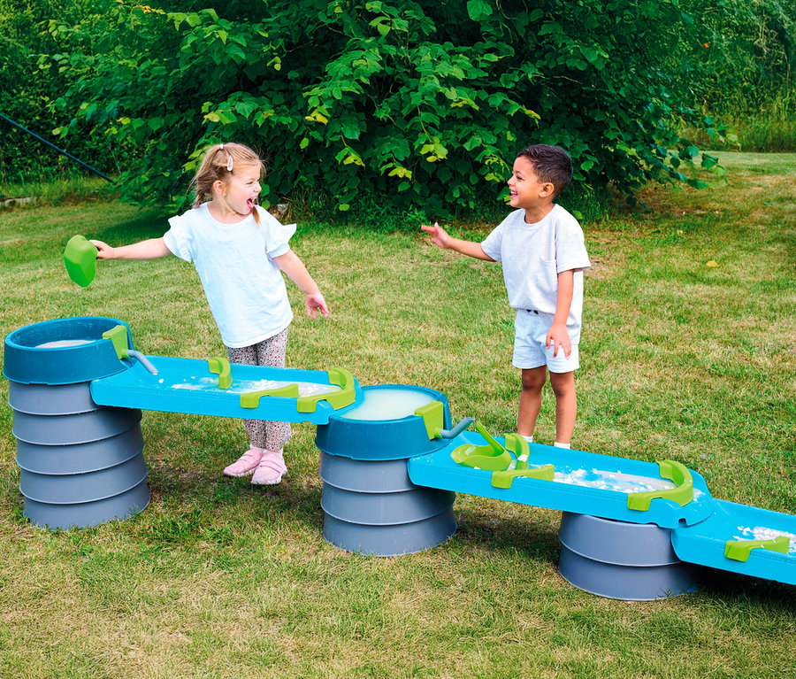 Zwei Kinder spielen mit der AquaPlay »Mudway STREAM« Matschbahn im Garten. Ein Mädchen gießt Wasser in die Bahn, ein Junge steht daneben.