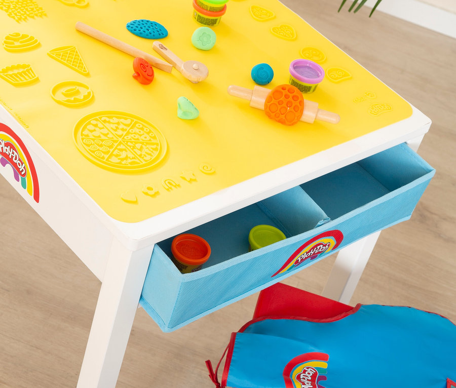 Auf einem Tisch mit gelber Spielmatte liegen Play-Doh-Dosen, Werkzeuge und Formen. In einer Schublade und einer Tasche befinden sich weitere Dosen.