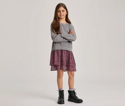 Ein junges Mädchen mit langen braunen Haaren steht mit verschränkten Armen und lächelt. Sie trägt ein graues Langarm-Sweatshirt, einen lila geblümten Stufenrock und schwarze Schnürstiefel mit hellvioletten Socken.