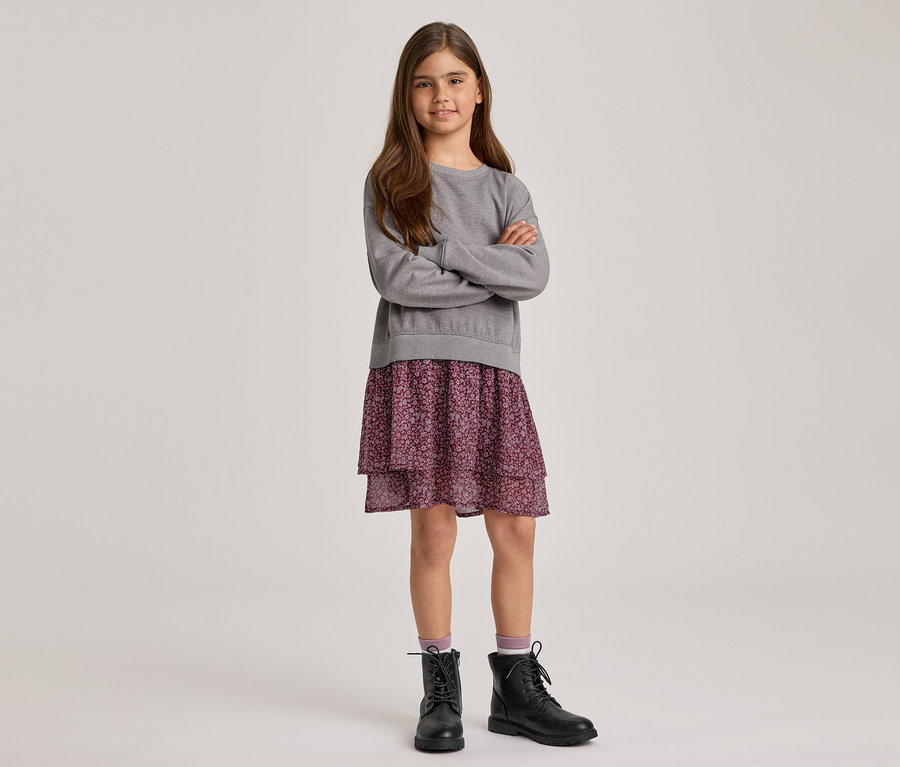 Ein junges Mädchen mit langen braunen Haaren steht mit verschränkten Armen und lächelt. Sie trägt ein graues Langarm-Sweatshirt, einen lila geblümten Stufenrock und schwarze Schnürstiefel mit hellvioletten Socken.