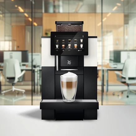 Ein Kaffeevollautomat mit Touchscreen, der Getränkeoptionen anzeigt, und einem Bohnenbehälter voller Kaffeebohnen. Ein Glas geschichteter Latte Macchiato steht auf der Abtropfschale, im Hintergrund ein verschwommenes Büro.