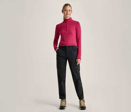 Eine Frau mit blondem Pferdeschwanz steht mit den Händen in den Taschen nach vorne gerichtet. Sie trägt ein pinkes Langarmshirt mit Reißverschluss am Hals, schwarze Hosen und braune Wanderschuhe.