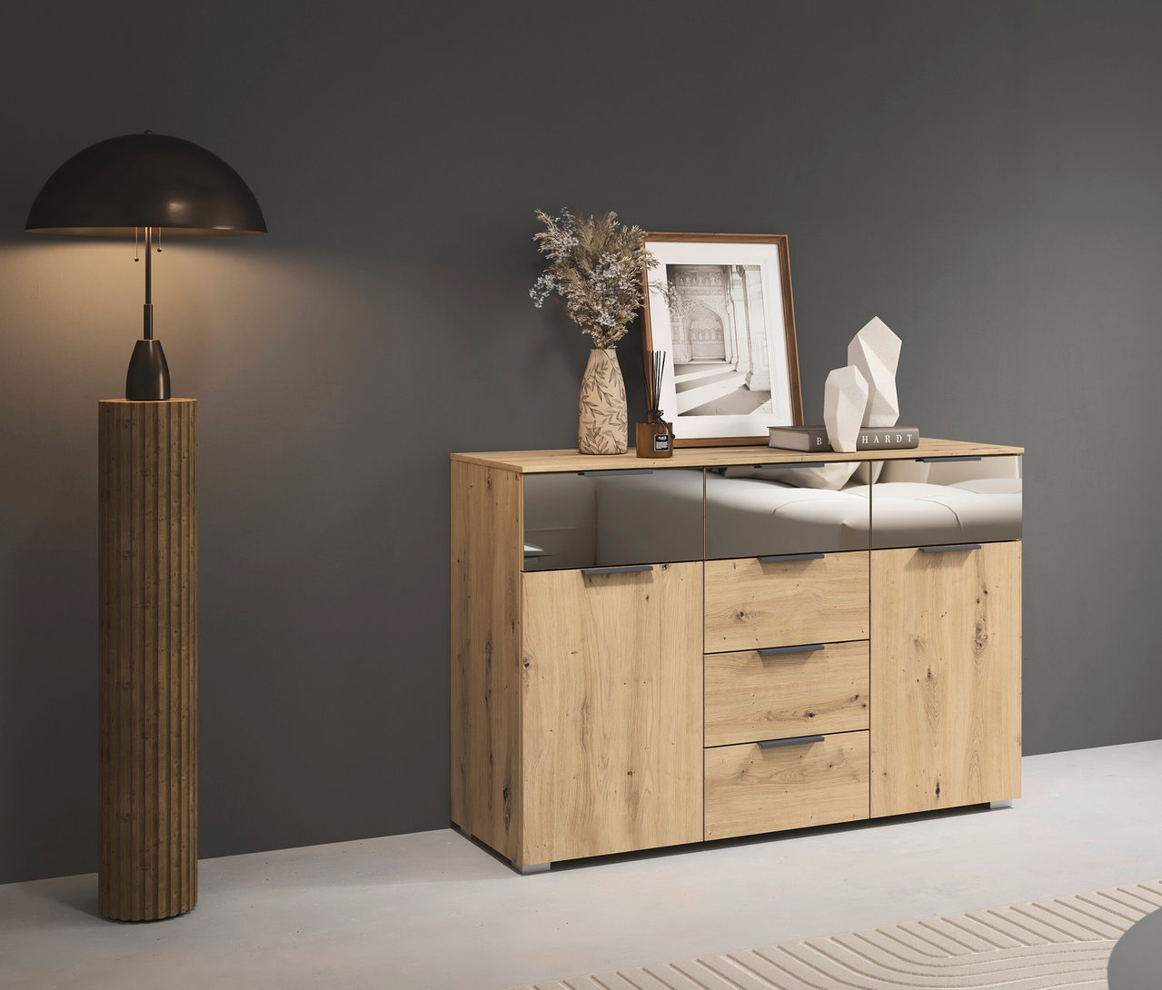 Eine rauch Kommode »Skyla« mit zwei Türen und sechs Schubladen in Eiche steht neben einer Stehlampe.