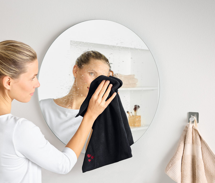 Eine Frau putzt einen beschlagenen runden Spiegel mit einem schwarzen Tuch.