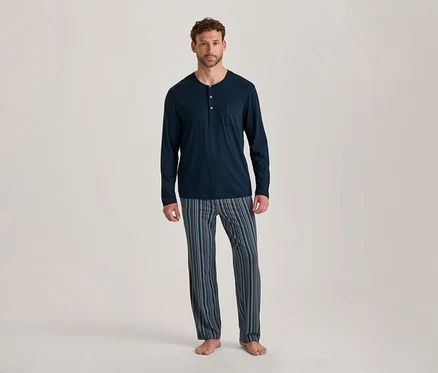 Mann steht barfuß, trägt ein dunkelblaues, langärmeliges Pyjama-Oberteil mit Knöpfen und Tasche sowie gestreifte Pyjamahosen in Blau, Braun und Schwarz.