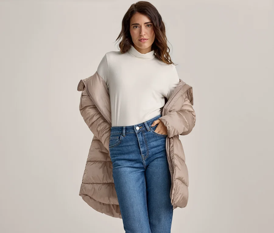 Frau mit dunklen Haaren trägt einen hellen Rollkragenpullover, blaue Jeans und eine beigefarbene Steppjacke über den Schultern, eine Hand in der Tasche, blickt in die Kamera.
