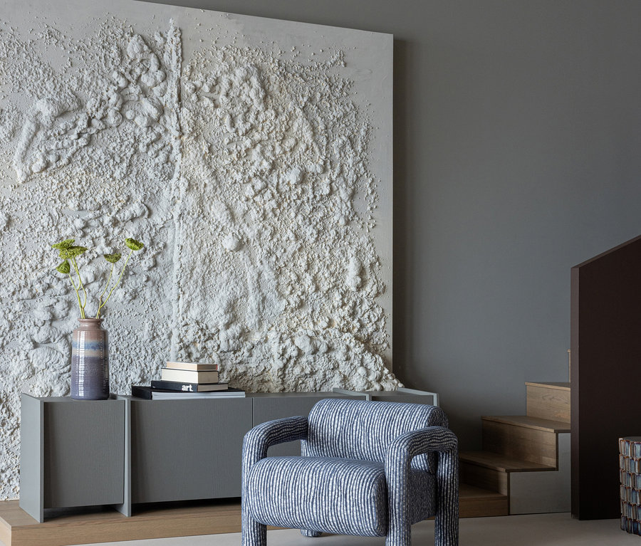 Ein moderner Raum zeigt ein großes, strukturiertes weißes Kunstwerk an einer grauen Wand. Darunter steht ein graues Sideboard mit einer Vase mit grünem Blattwerk und einem Stapel Bücher. Ein blau-weiß gestreifter Sessel steht davor. Rechts ist eine Holztreppe sichtbar.