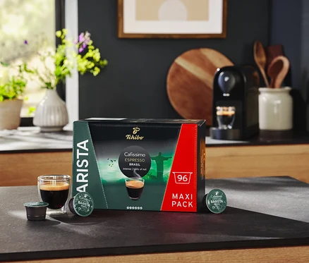 Auf einem Tisch stehen eine Packung Cafissimo BARISTA Espresso Brasil - 96 Kapsel, ein Glas Espresso und eine Kaffeemaschine.