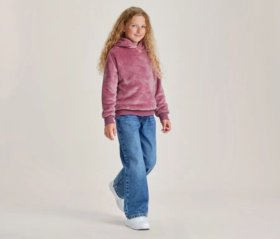Ein junges Mädchen mit langen, lockigen blonden Haaren lächelt und geht vorwärts, trägt einen flauschigen pinken Hoodie, blaue Jeans und weiße Sneaker.