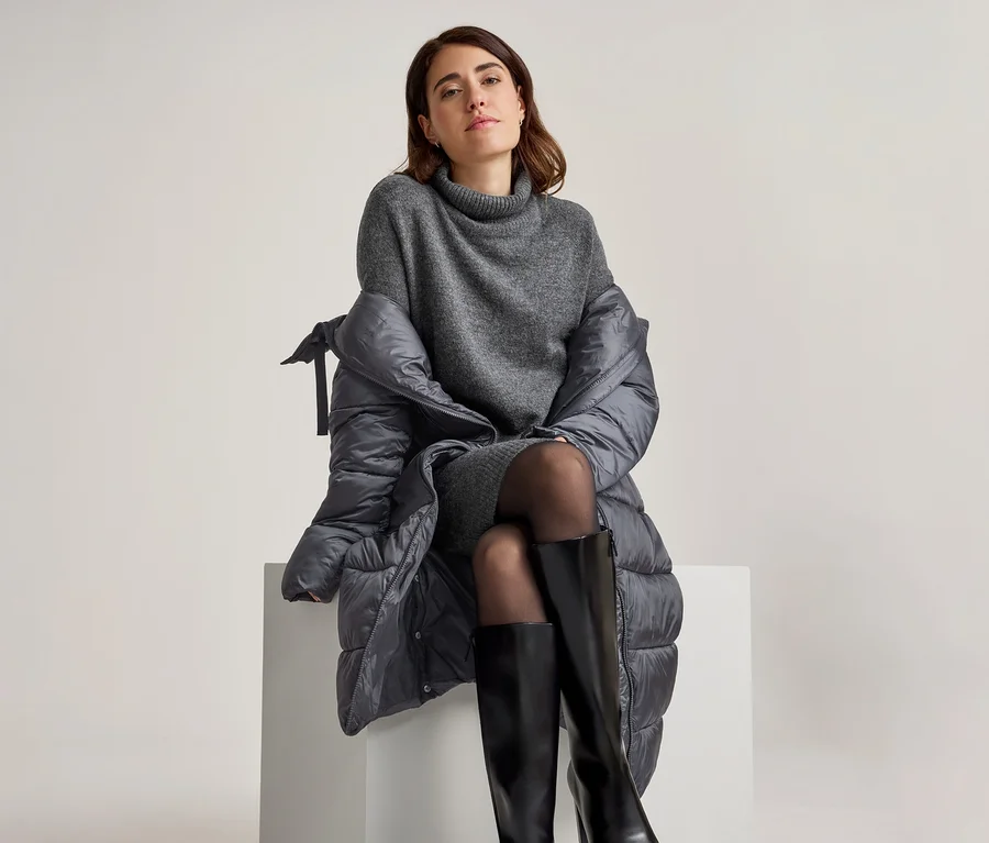 Frau sitzt auf einem weißen Block, trägt ein graues Rollkragenkleid, schwarze Strumpfhosen und schwarze kniehohe Stiefel, mit einem dunkelgrauen Steppmantel über den Schultern.