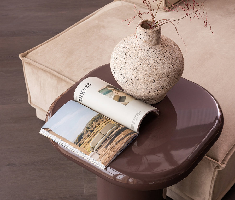 Brauner Beistelltisch mit offenem Magazin und strukturierter Vase mit getrocknetem Gras, neben einem beigen Sofa auf dunklem Holzboden.