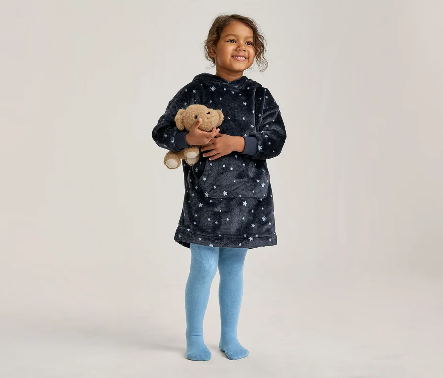 Junges Mädchen hält einen Teddybär. Sie trägt einen dunkelblauen, flauschigen Kapuzenpullover mit Sternen und hellblaue Strumpfhosen.