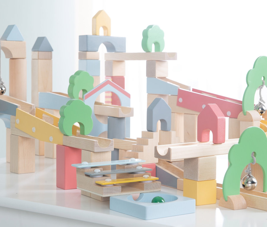 Ein komplexes Holz-Kugelbahn- und Baustein-Set mit Rampen, Bögen und pastellfarbenen Blöcken in Blau, Rosa, Gelb und Grün. Kleine Holzbäume, Häuser und Glocken sind integriert, mit einer grünen Kugel in einer blauen Basis.