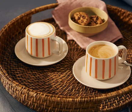 Zwei weiße Kaffeetassen mit orangen Streifen, eine mit Milchschaum, die andere mit Kaffee, stehen mit Keksen und einer rosa Serviette auf einem geflochtenen Tablett.