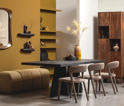 Modernes Esszimmer mit dunklem Holztisch, drei beigefarbenen Stühlen und einer passenden Bank. Eine senfgelbe Wand hat einen Holzspiegel und zwei Regale mit Büsten. Ein hoher Holzschrank steht daneben, eine Vase mit Pampasgras auf dem Tisch.