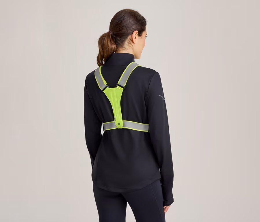 Frau von hinten, trägt ein schwarzes Langarmshirt und schwarze Leggings, darüber eine neongelbe Reflektorweste mit reflektierenden Streifen und einem kleinen Lichtknopf.