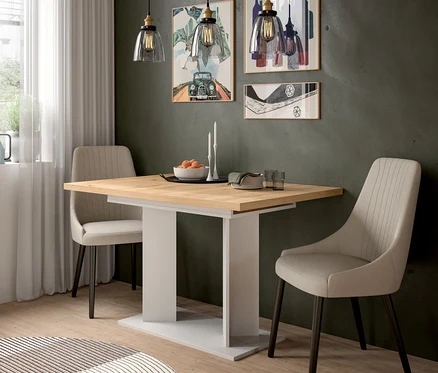 Ein moderner Essbereich mit einem hellen Holztisch auf weißem Untergestell, flankiert von zwei beigefarbenen Polsterstühlen mit dunklen Beinen. Drei Pendelleuchten mit klaren Glasschirmen beleuchten den Tisch, auf dem eine Obstschale, weiße Kerzen, gestapelte Teller und Gläser stehen. Drei gerahmte Bilder schmücken die dunkle Wand, und ein runder gemusterter Teppich liegt auf dem Holzboden.