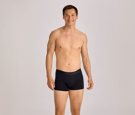 Oberkörperfreier Mann in schwarzen Boxershorts lächelt in die Kamera.