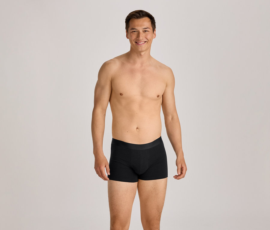 Oberkörperfreier Mann in schwarzen Boxershorts lächelt in die Kamera.