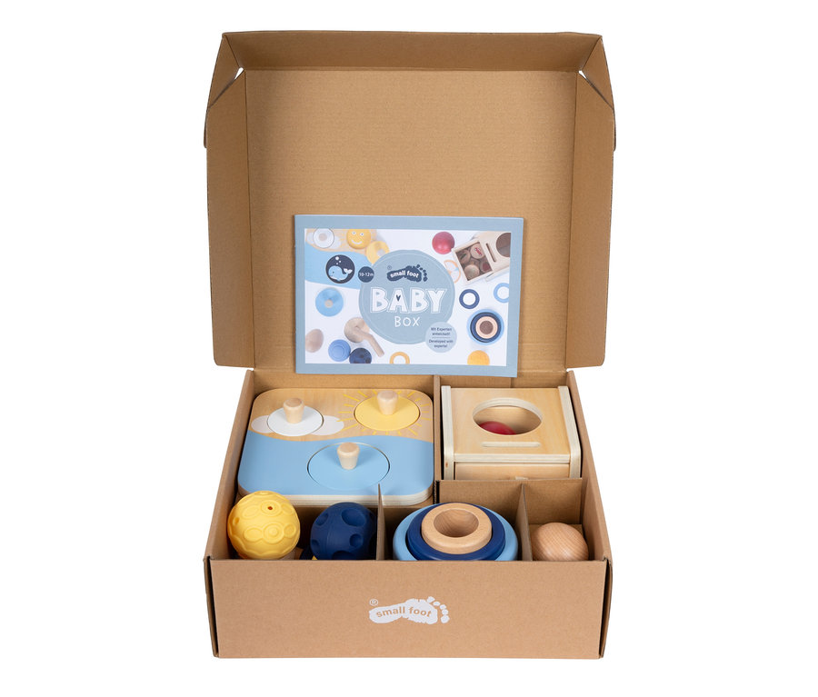 Offene braune Kartonbox mit einem "BABY BOX"-Heft und verschiedenen Holz- und Fühlspielzeugen für Babys, darunter ein Formensortierer, Stapelringe und Bälle.