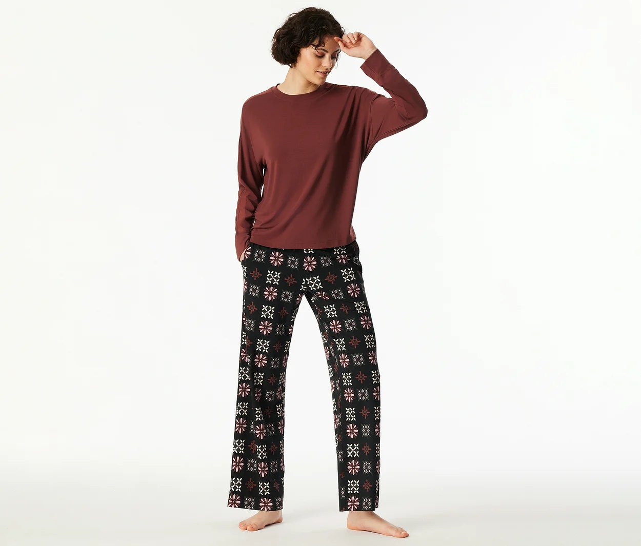Frau mit kurzen Haaren trägt ein rotbraunes Langarmshirt und eine schwarze Pyjamahose mit rosa-weißem Blumenmuster, barfuß stehend.
