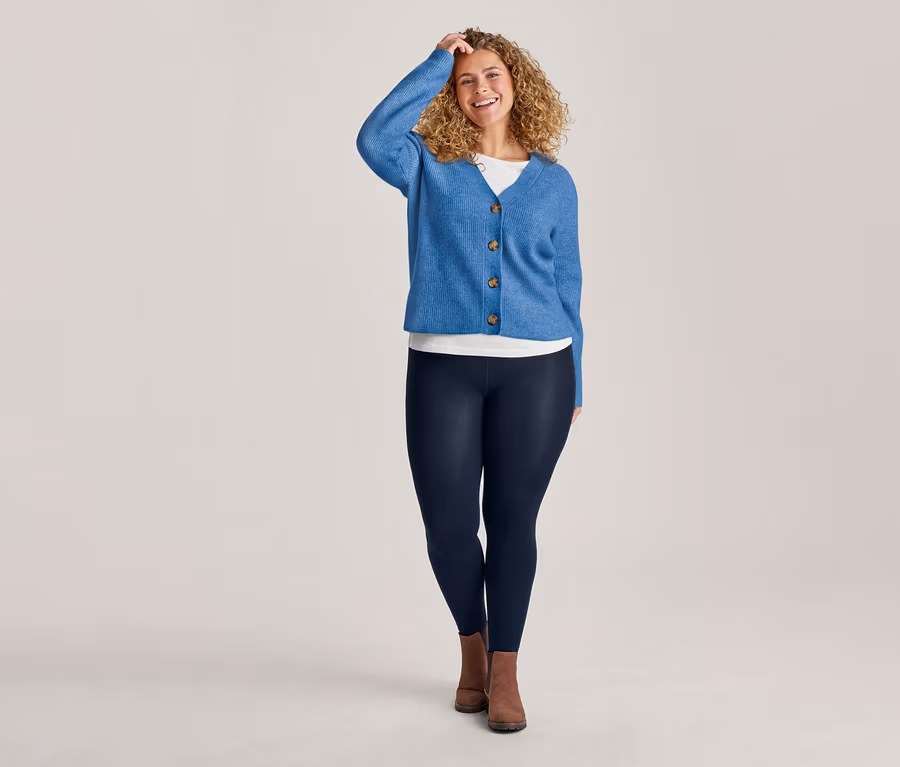 Lächelnde Frau mit lockigem Haar trägt eine blaue Strickjacke mit RWS-zertifizierter Wolle über einem weißen Oberteil und dunkelblaue Thermoleggings.