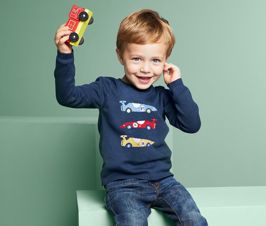 Ein Junge sitzt und hält ein hölzernes Spielzeugauto hoch. Er trägt einen blauen Feinstrick-Pullover mit Auto-Motiven und Jeans.