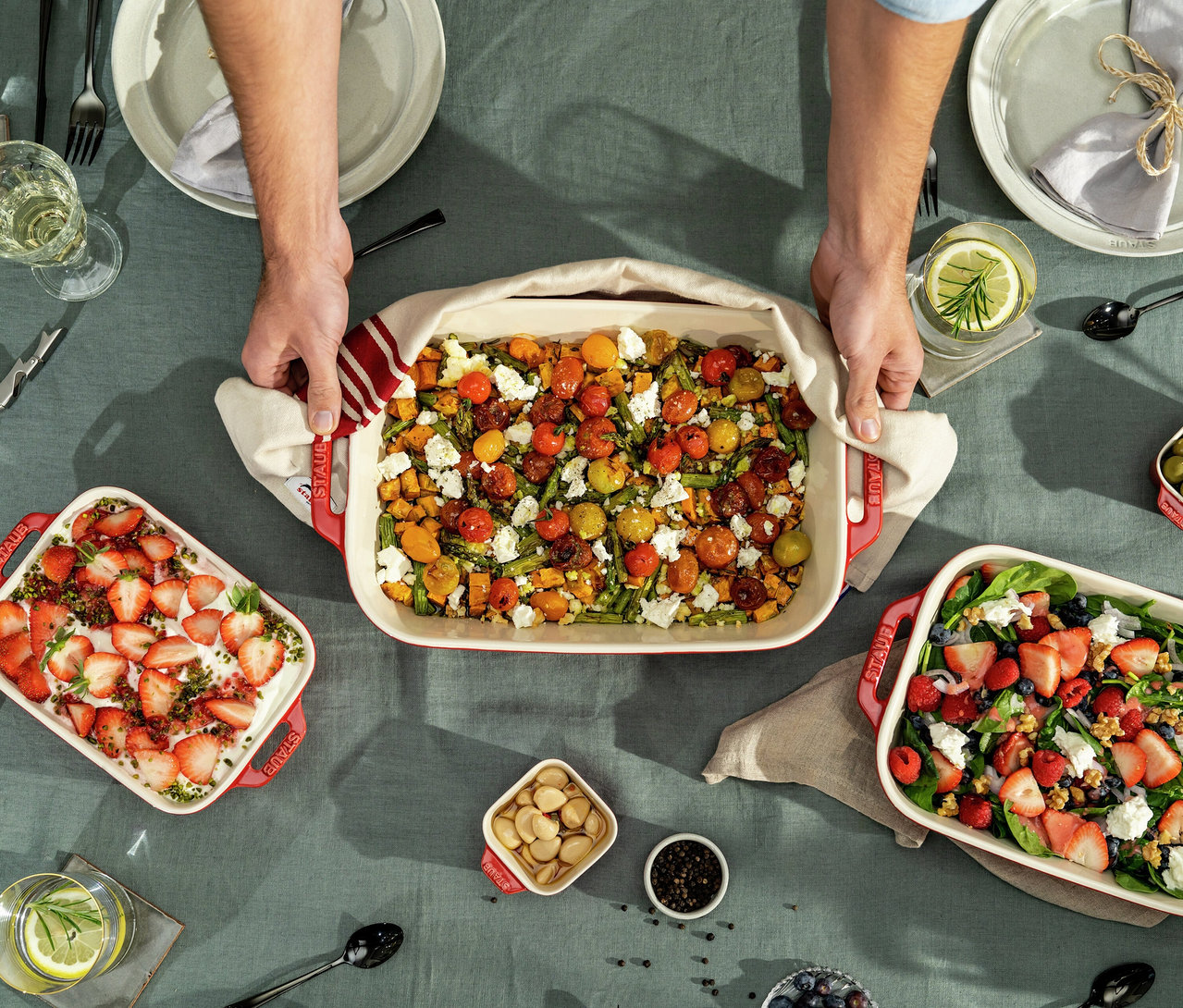 Hände stellen eine große kirschrote STAUB Auflaufform mit geröstetem Gemüse, Tomaten, Spargel und Feta auf einen dunkelgrünen Tisch. Zwei weitere kirschrote STAUB Auflaufformen stehen auf dem Tisch: eine mit Erdbeeren und Sahne, die andere mit einem Beeren-Spinat-Salat. Der Tisch ist mit Tellern, Gläsern und kleinen Schalen mit Nüssen gedeckt.