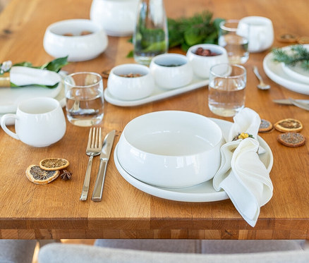 Ein gedeckter Holztisch mit weißem Geschirr. Eine weiße Schale des Mäser Teller-Sets »Taya« steht auf einem Teller, daneben eine Serviette, Besteck, eine Tasse und ein Wasserglas. Im Hintergrund weitere Schalen und eine Flasche.