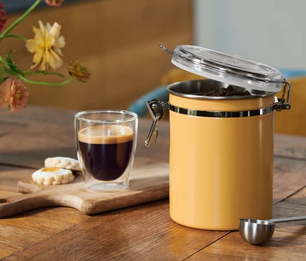 Kaffee in einem Glas, Kekse auf einem Holzbrett und ein offener gelber Kaffeebehälter mit Bohnen, dazu ein Messlöffel, auf einem Holztisch mit Blumen.