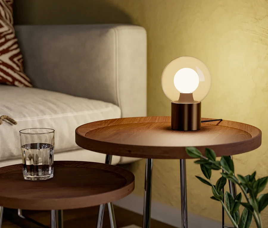 Ein modernes Wohnzimmer mit zwei runden Beistelltischen aus Holz. Eine kugelförmige Tischlampe mit bernsteinfarbenem Schirm leuchtet auf dem größeren Tisch, neben einem hellgrauen Sofa mit einem rot-weiß gemusterten Kissen. Ein Glas Wasser steht auf dem kleineren Tisch, und ein geometrischer Teppich bedeckt den Boden.