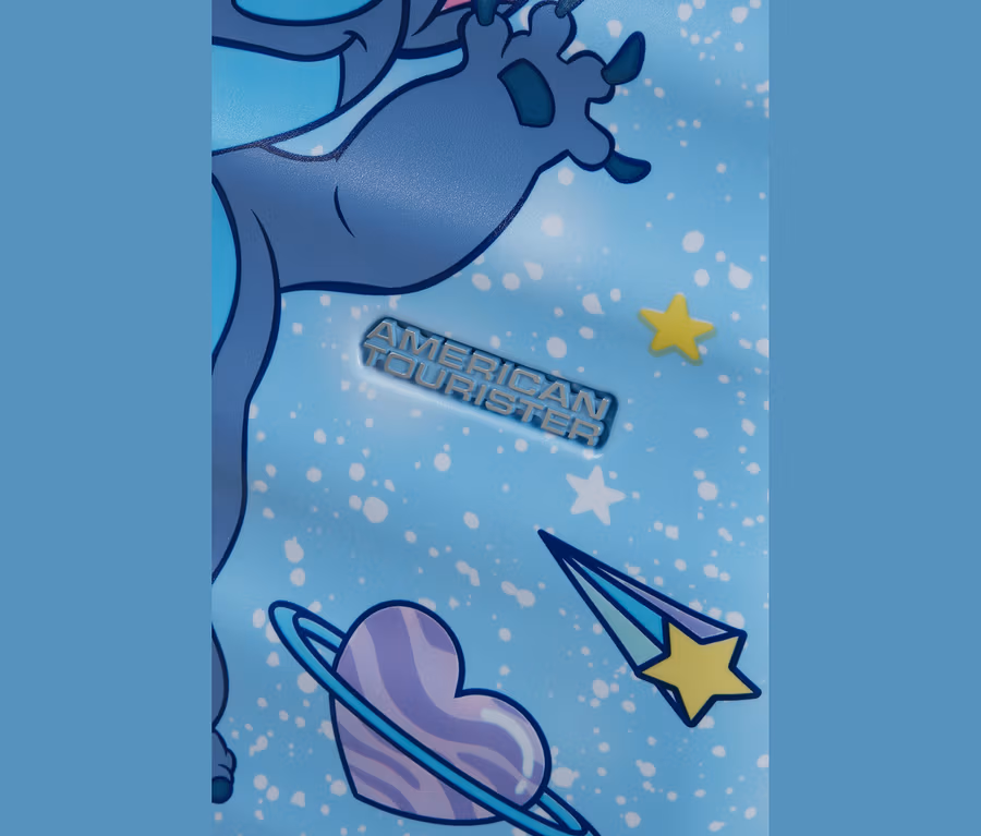 Nahaufnahme einer hellblauen Oberfläche mit weißen Sprenkeln, die einen Teil der blauen Zeichentrickfigur Stitch mit Krallen, das silberne American Tourister Logo, einen herzförmigen Planeten, einen gelben Stern und eine Sternschnuppe zeigt.