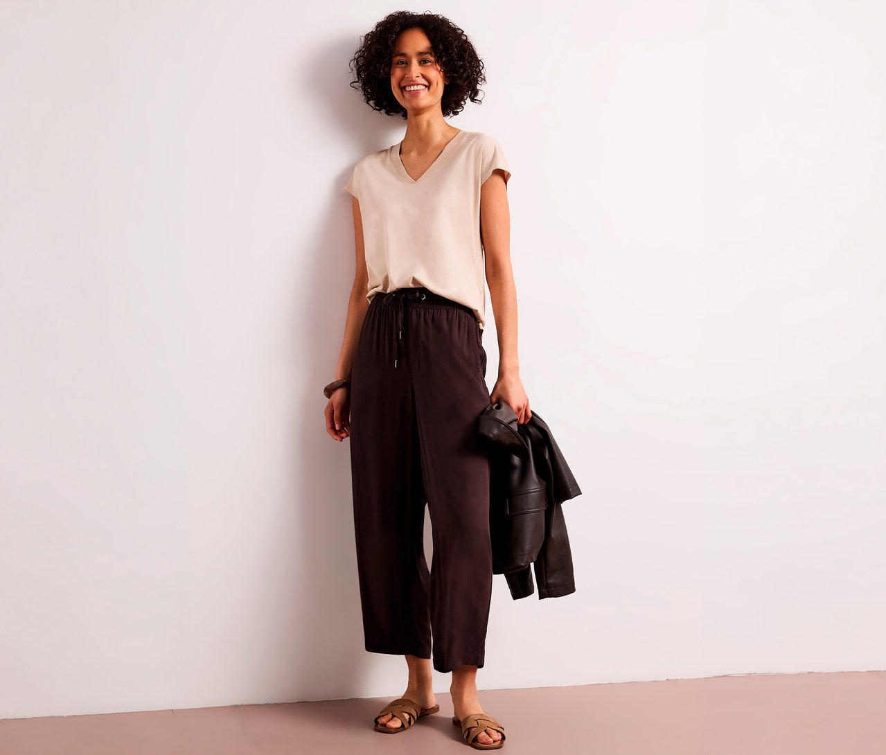 Lächelnde Frau mit lockigem Haar trägt beiges V-Neck-Top und braune Street One 7/8-Culotte, hält eine Jacke, steht an einer weißen Wand.
