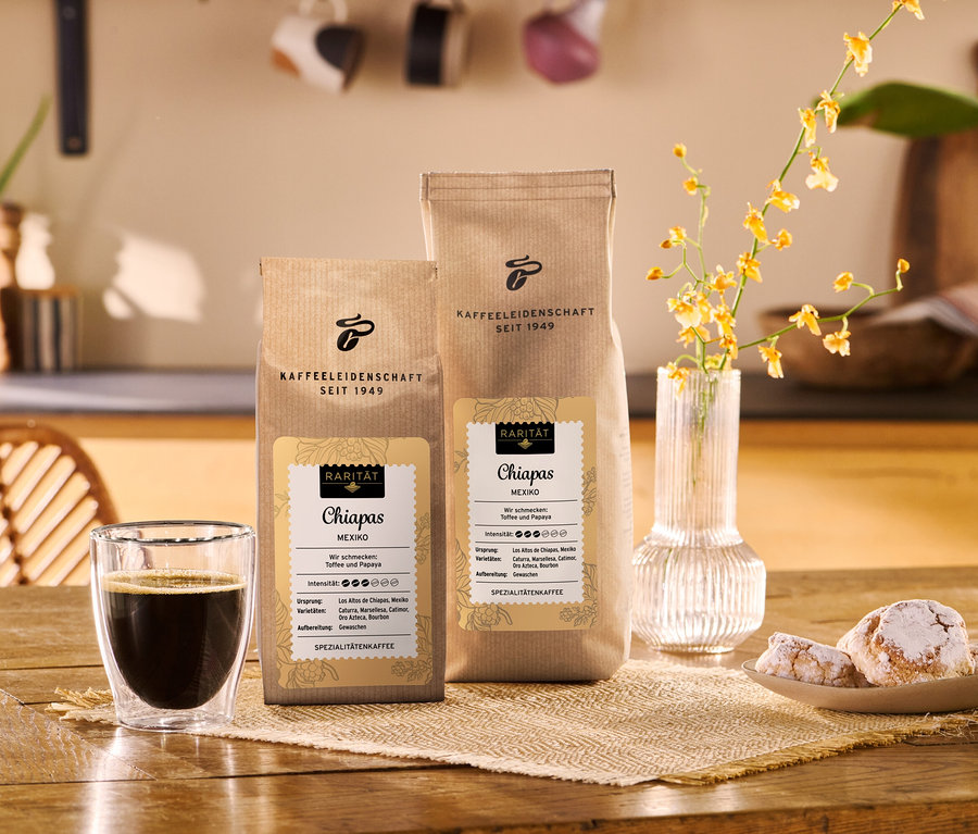 Zwei braune Kaffeepackungen mit der Aufschrift „Chiapas Mexiko“ stehen auf einem Holztisch neben einem Glas schwarzem Kaffee, einer Vase mit gelben Blumen und einem Teller mit Puderzuckerkeksen.