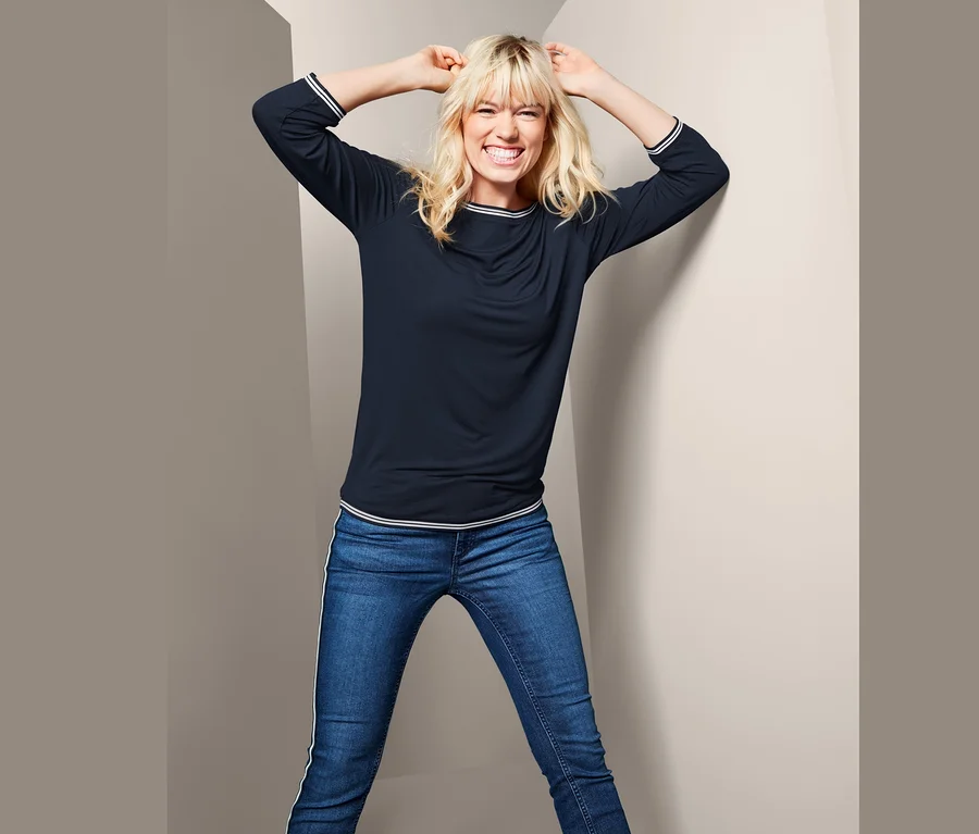 Frau lächelt und posiert in einem Blusenshirt mit 3/4-Arm und Jeans.