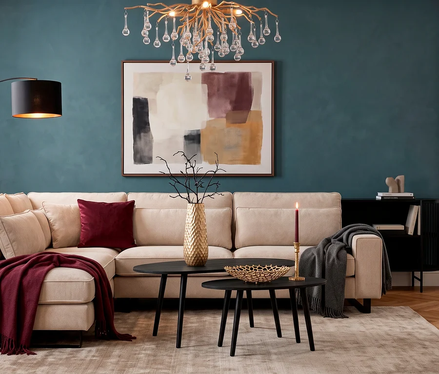 Ein beiges Ecksofa »Veluno« mit einer pflaumefarbenen SCHÖNER WOHNEN-Kollektion Wohndecke »BASICS« und einem burgunderroten Dekokissenbezug »SOUL« steht vor einer petrolfarbenen Wand. Davor sind zwei schwarze Eglo Tische »ALORAN« mit einer goldenen Vase »NILGAUT«, einer Schale »Kimitsu« und einem Kerzenhalter »JELLICOE« platziert. Eine Messing-Deckenleuchte und eine Trio Stehleuchte »Bolivia« beleuchten den Raum.