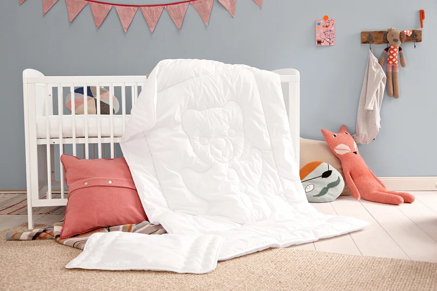 Weiße Steppdecke mit Kissen und rosa Dekokissen neben einem weißen Kinderbett.