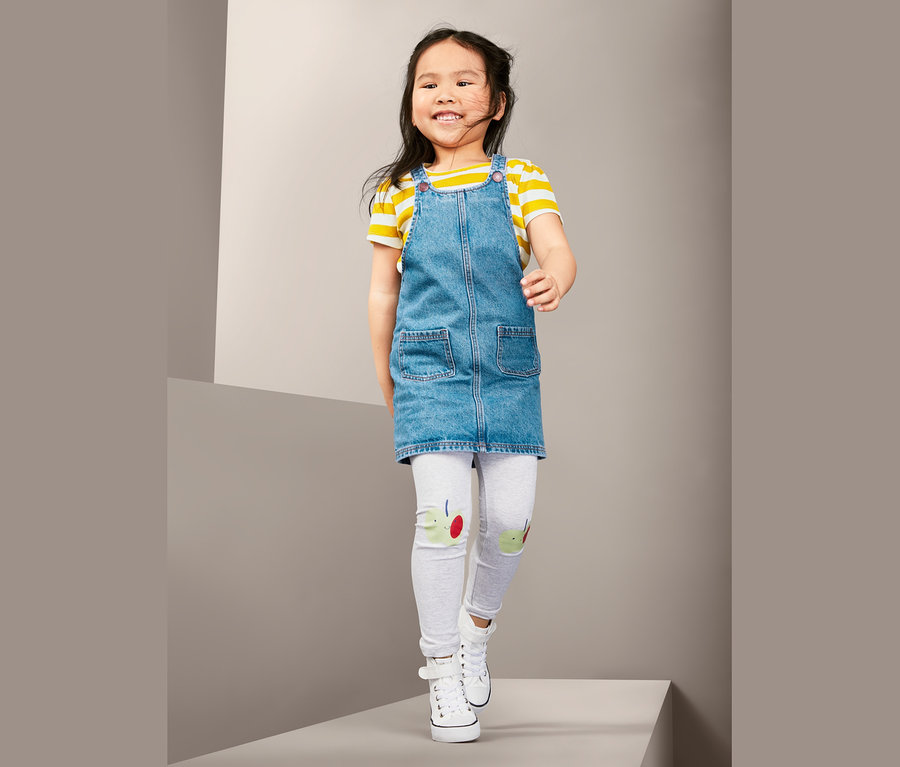 Lächelndes Mädchen in Denimlatzkleid, gelb-weiß gestreiftem T-Shirt, grauen Leggings und weißen Turnschuhen geht auf einer grauen Fläche.