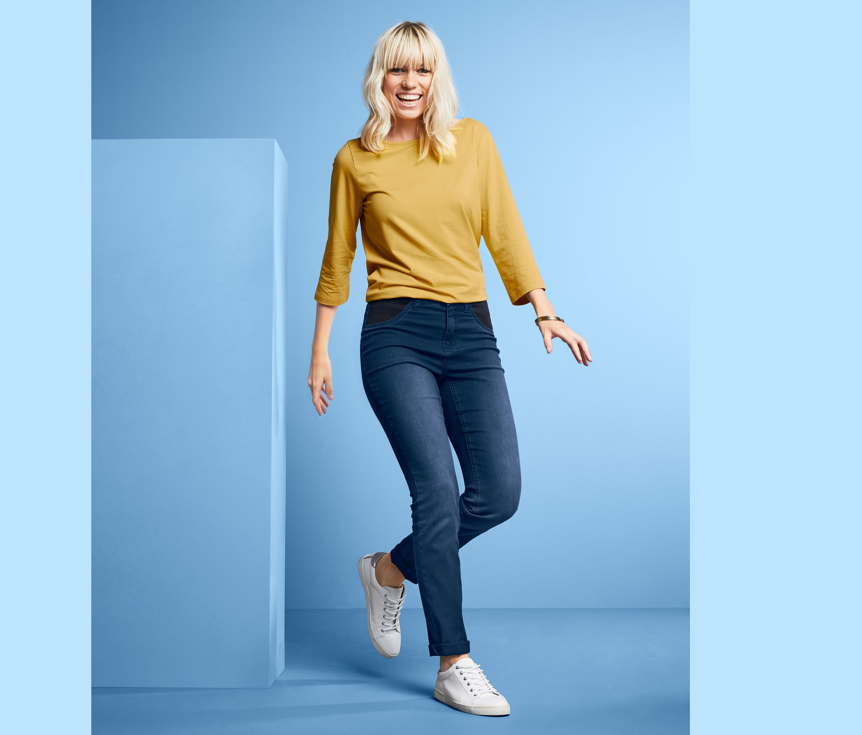 Blonde Frau posiert in Jeans mit Komfortbund, gelbem Oberteil und weißen Schuhen vor blauem Hintergrund.