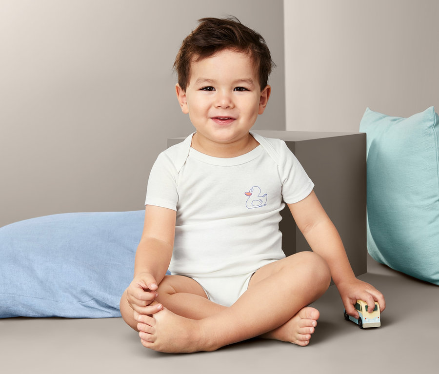 Ein kleiner Junge sitzt mit gekreuzten Beinen auf dem Boden und trägt einen Baby-Body. Im Hintergrund sind zwei Kissen zu sehen.