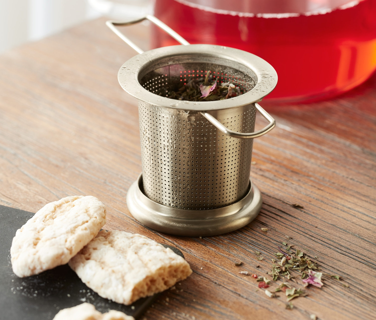 Ein Teesieb aus Edelstahl mit Kräutern steht auf einem Holztisch neben Keksen und einem Krug mit rotem Getränk.