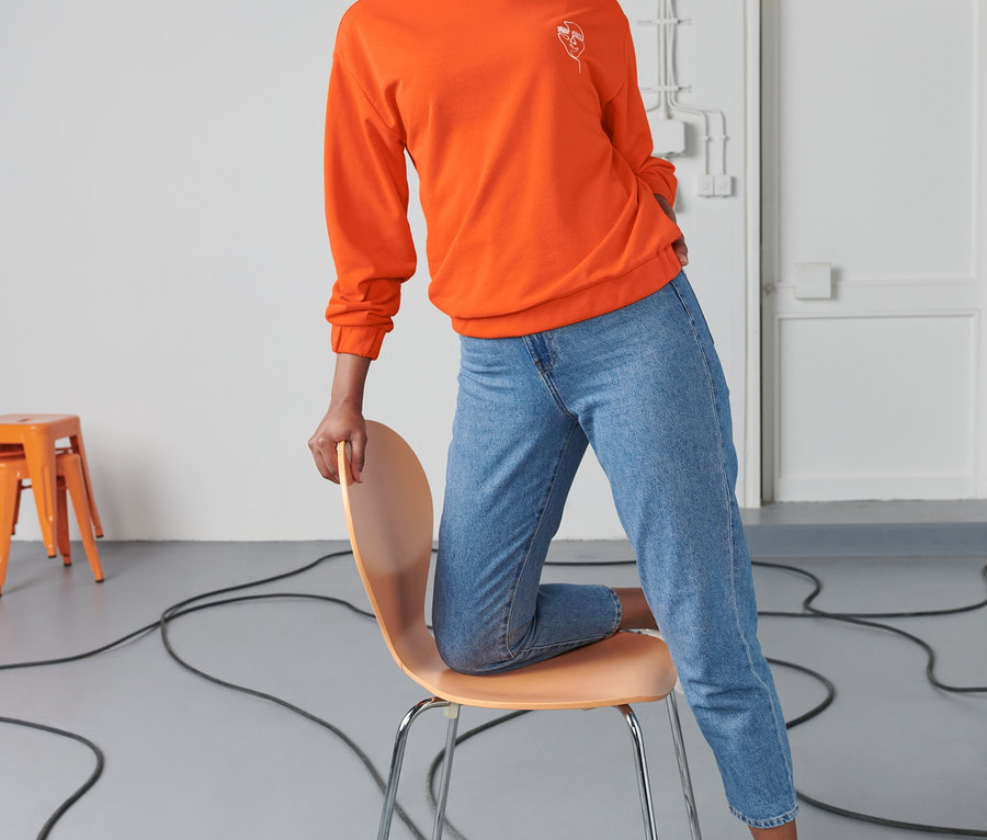 Frau sitzt auf einem Stuhl und trägt ein orangefarbenes Sweatshirt mit Stickerei.