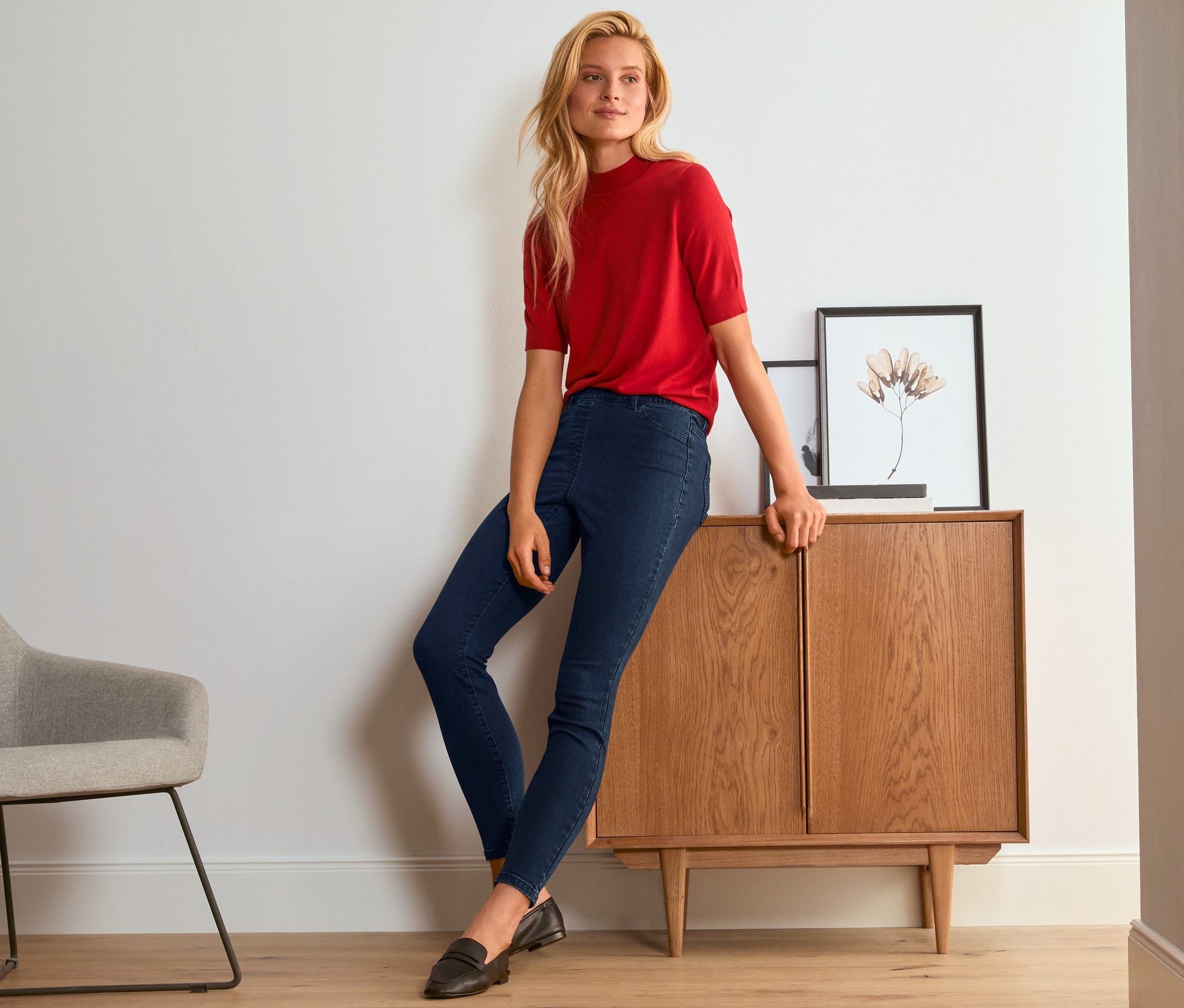 Blonde Frau in rotem Pullover und blauen Treggings lehnt an einem Holzschrank.