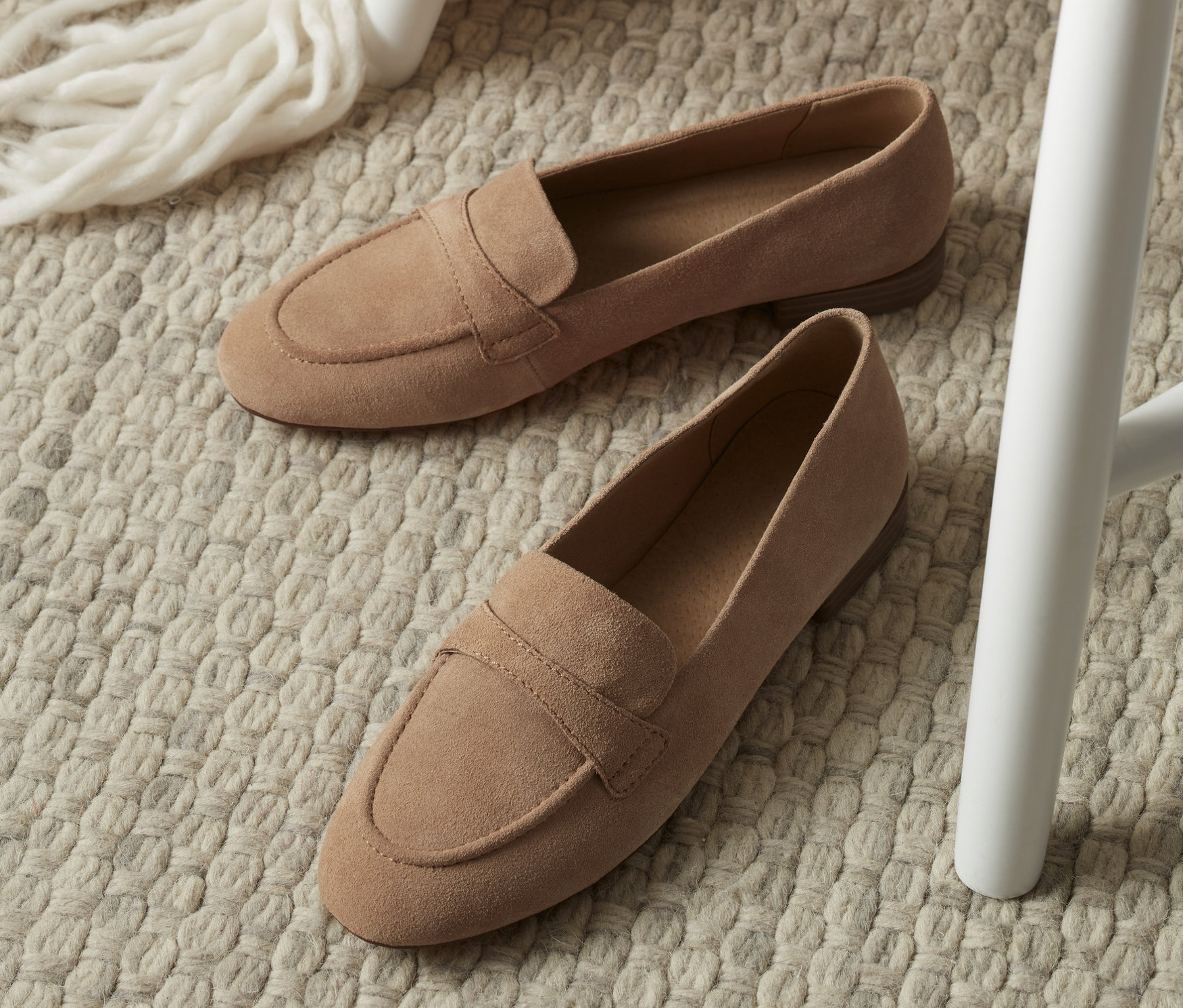 Zwei Loafer aus Veloursleder liegen auf einem beigen Teppich neben einem weißen Stuhl.