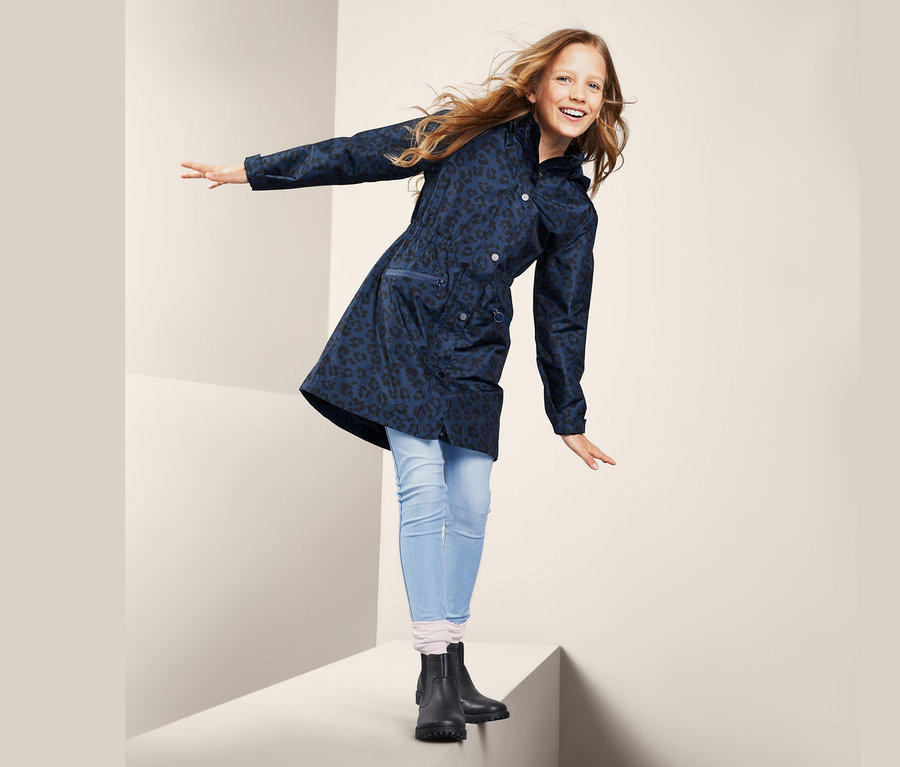 Mädchen steht auf einem weißen Würfel und winkt mit den Händen. Sie trägt einen Allwetterparka mit Alloverprint, Jeans und schwarze Stiefel.