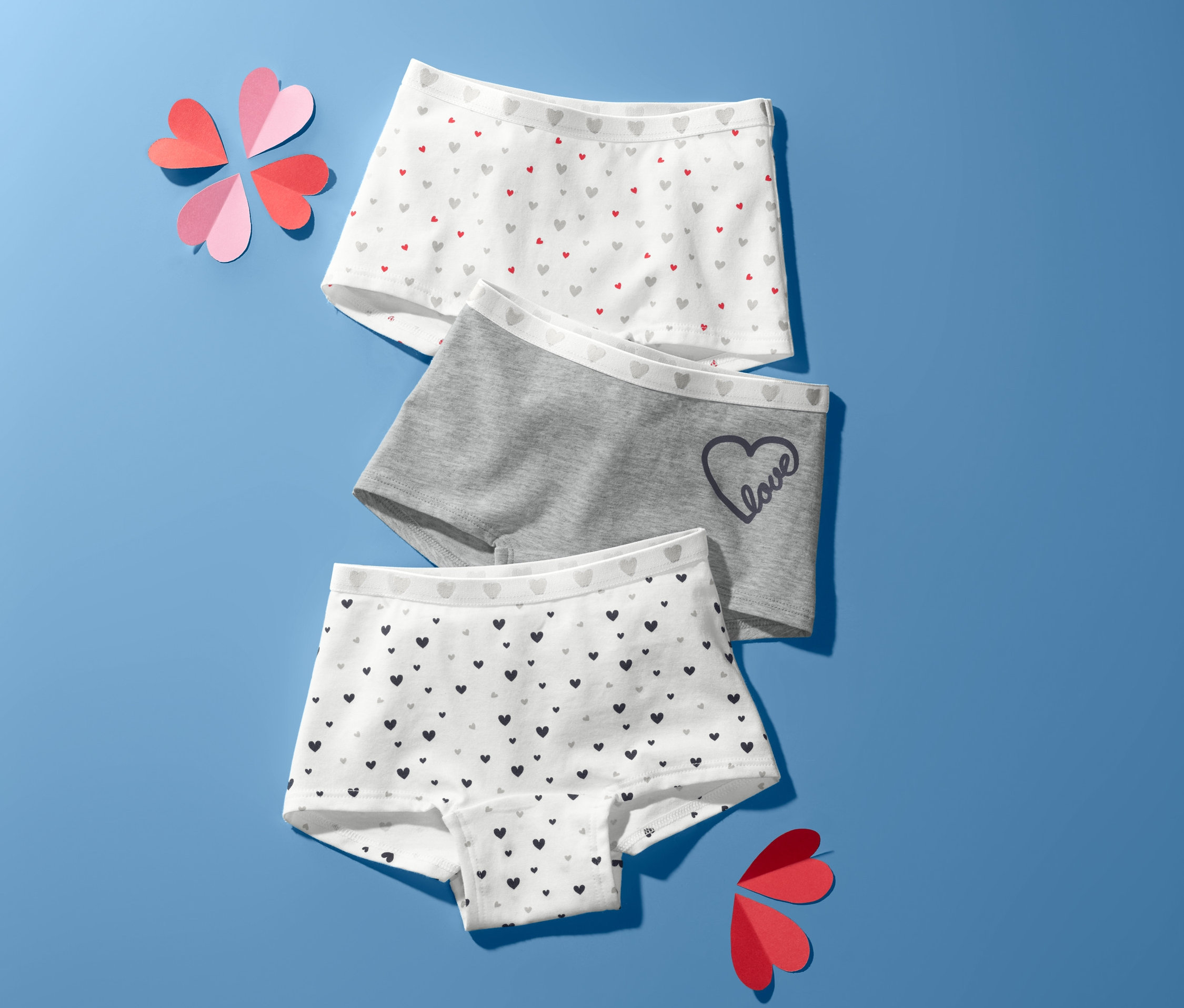 Drei Mädchen-Pantys liegen auf blauem Hintergrund, zusammen mit herzförmigen Dekorationen.